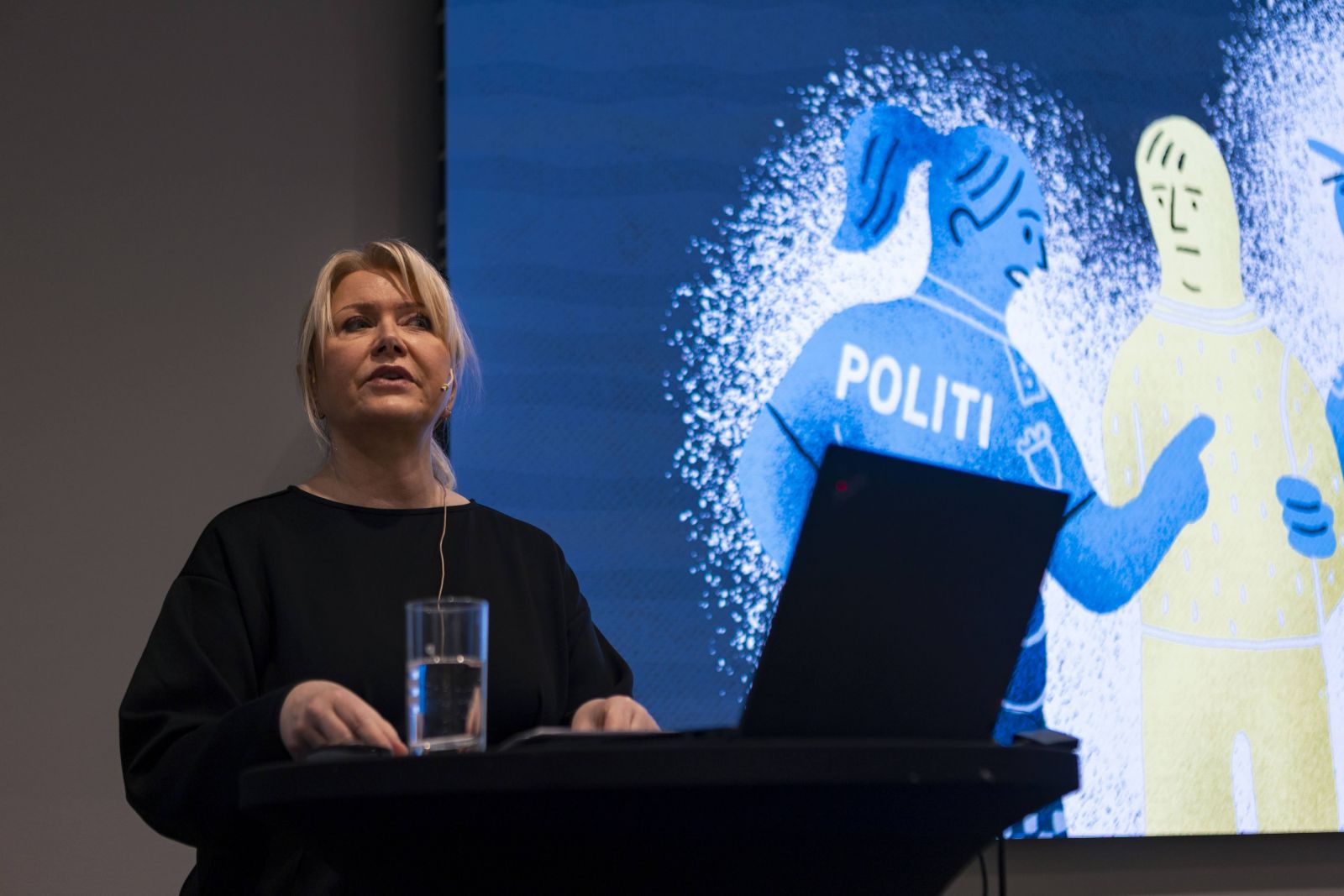 Bildet viser en dame som står på scenen og snakker. Lysbilde i bakgrunnen.