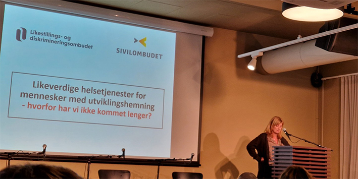 Bilde av en dame som står ved talerstol, bak henne er det et lerrett med tittel likeverdig helsetjenester for mennesker med utviklingshemming - hvorfor har vi ikke kommet lengre 