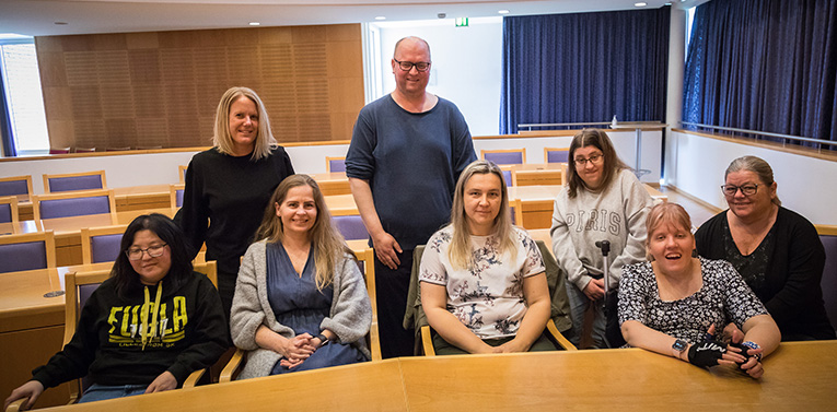 8 personer sitter i et klasserom. Alle ser mot kameraet. 