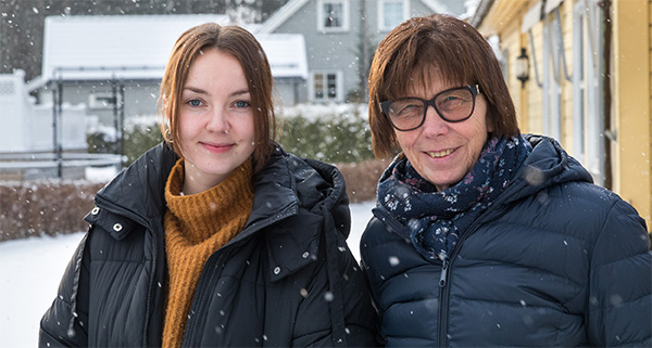 To damer står utendørs. Det snør. De ser mot kameraet og smiler. 